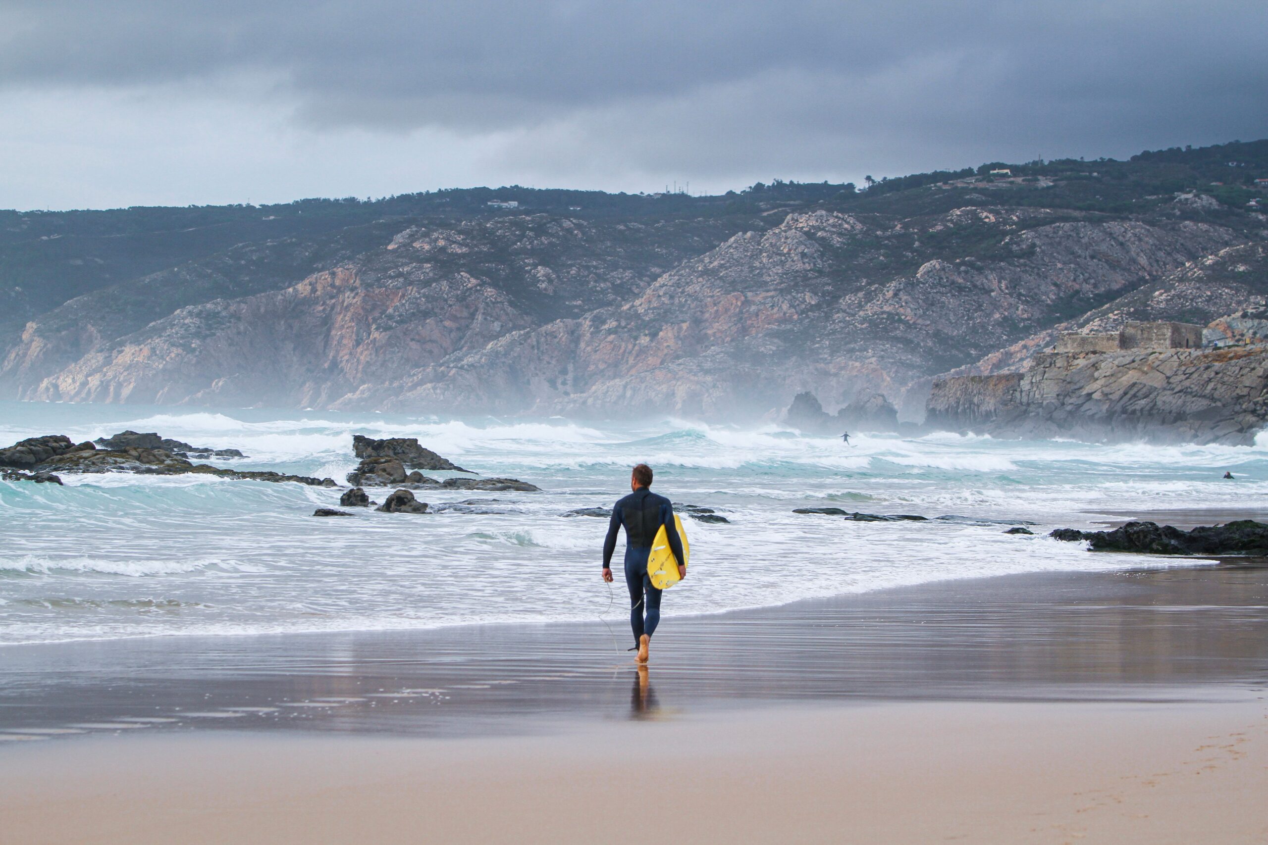 A male surfer walking on a sandy beach towards the ocean carrying a surfboard under his arm.