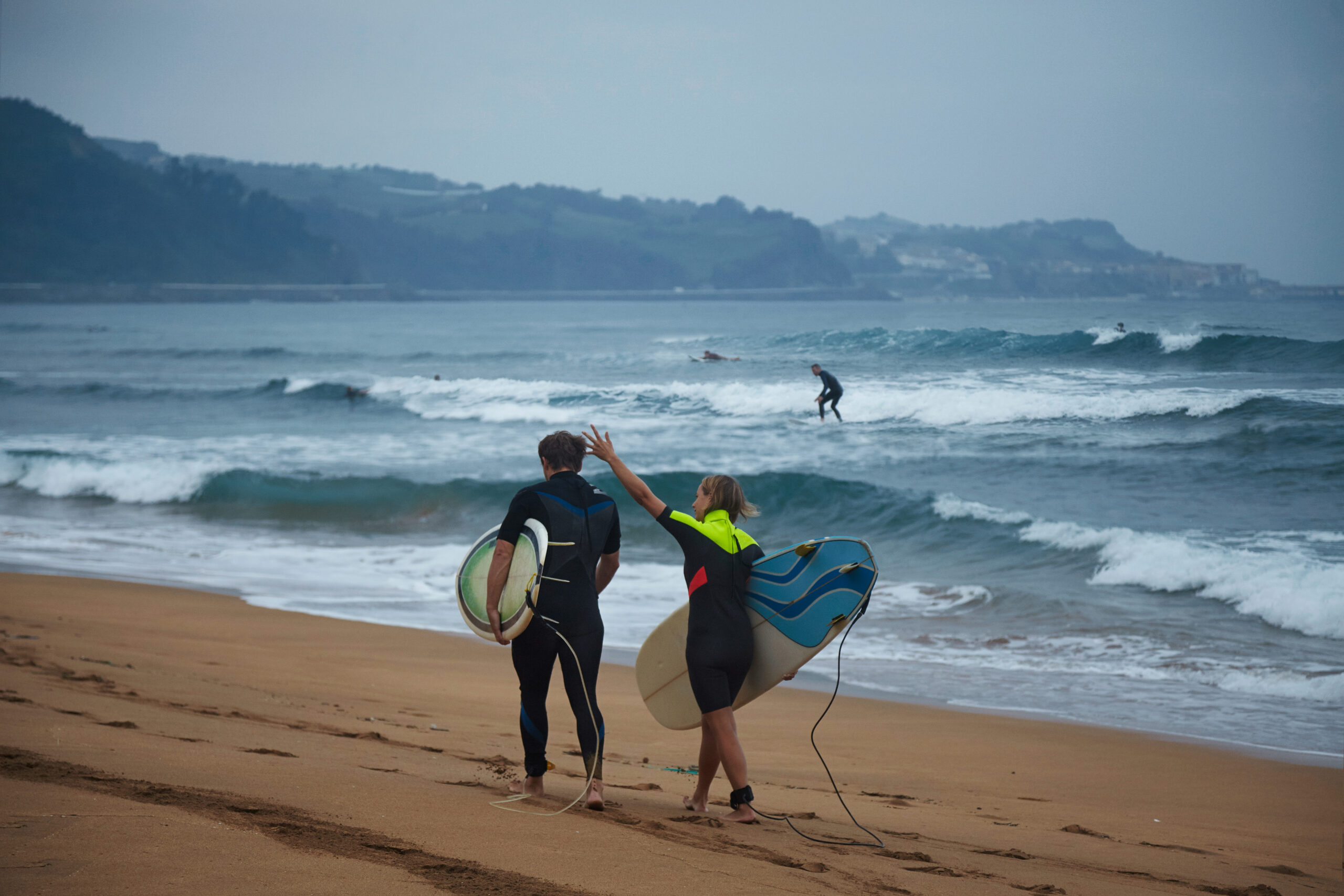 Surf couple walks and talks, carrying their longboards on ocean beach shore after surfing, professional surfer caught the wave on background